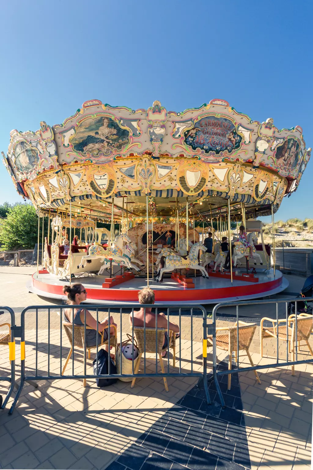 A vintage carousel with colorful horses spins in the sunlight, while several people sit in chairs in front of it.