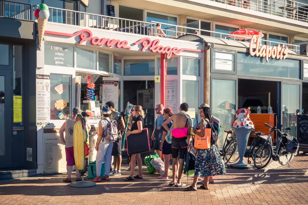 A line of people waits outside an ice cream shop. Some guests wear swimwear, while others carry bags.