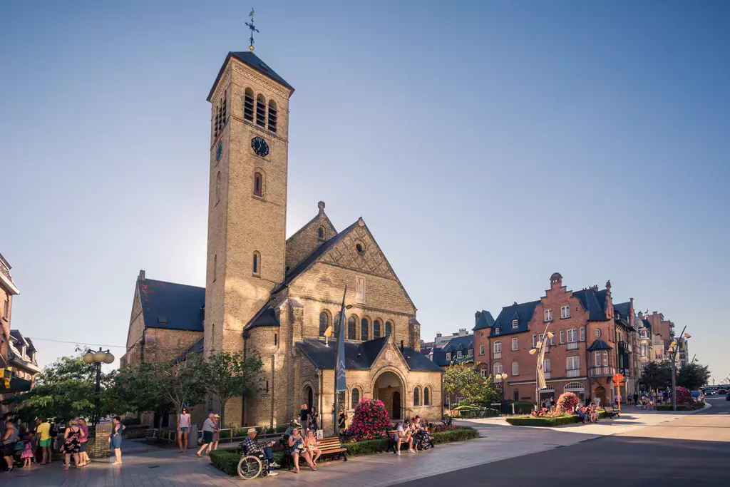 An old church with a tall tower stands at a square, surrounded by blooming flowers and several pedestrians.