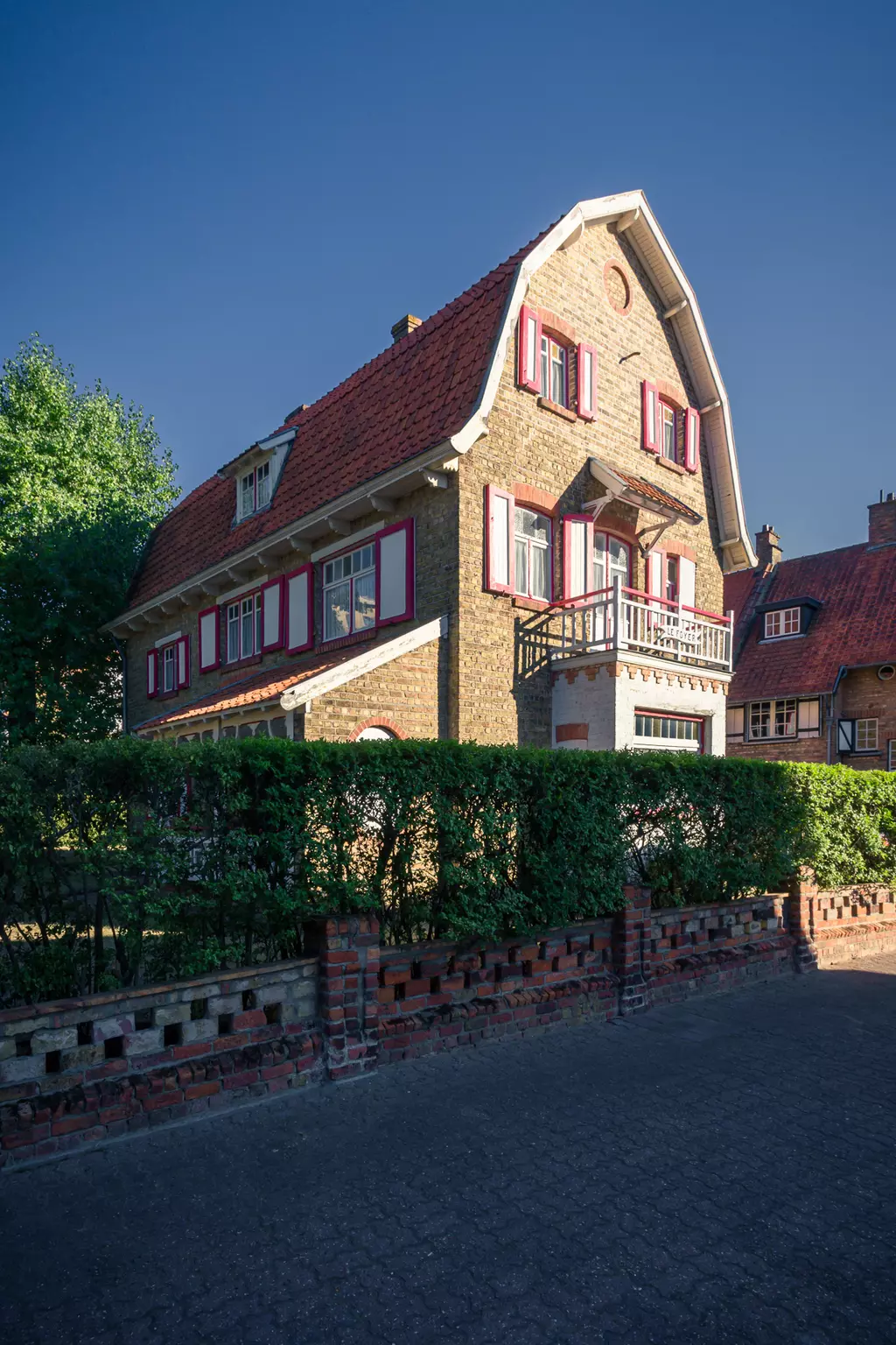 A three-story house with a red tiled roof, white windows, and a small balcony, surrounded by a well-maintained garden.