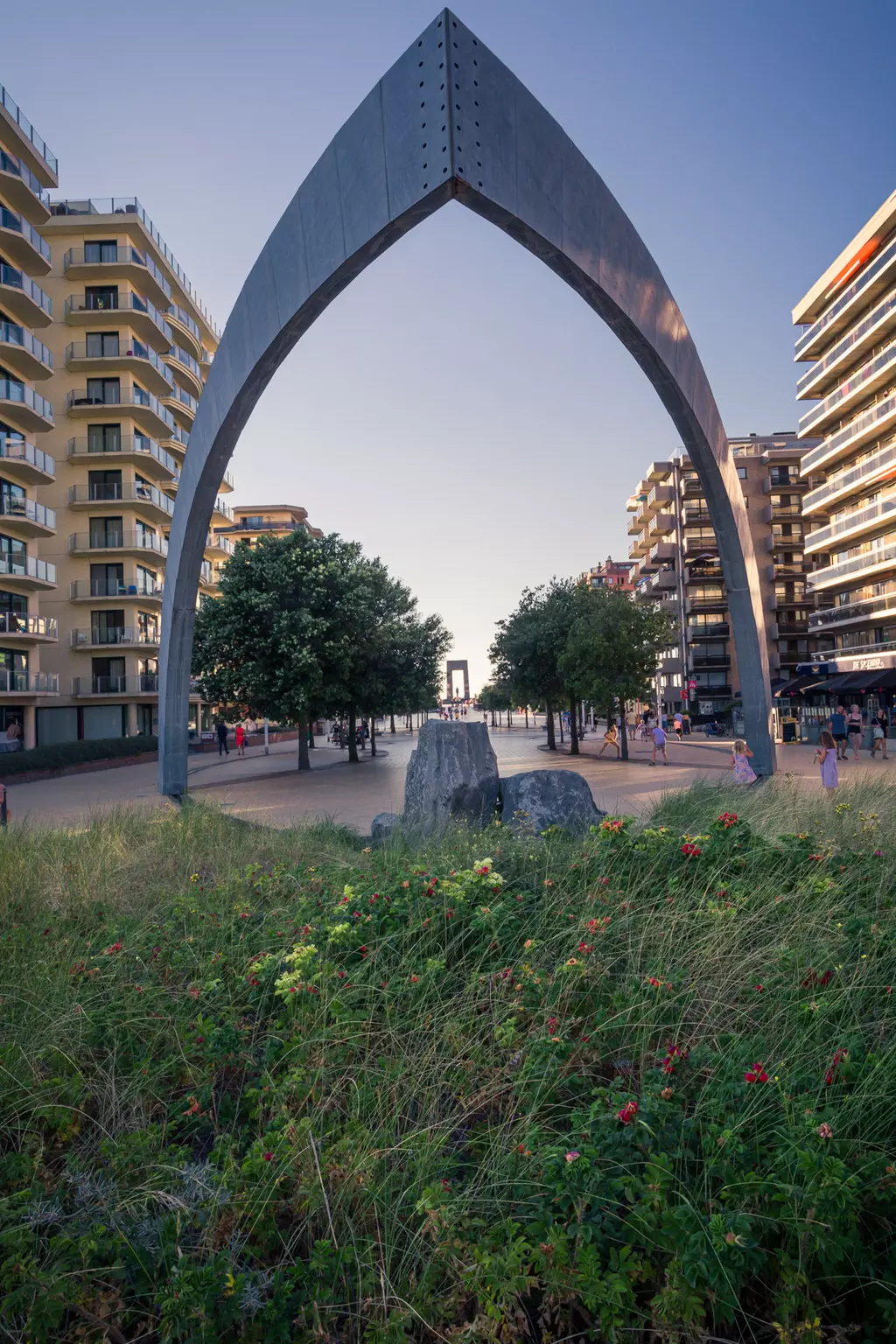 A large, arch-shaped metal sculpture stands in the foreground, surrounded by greenery and flowers, with residential buildings in the background.