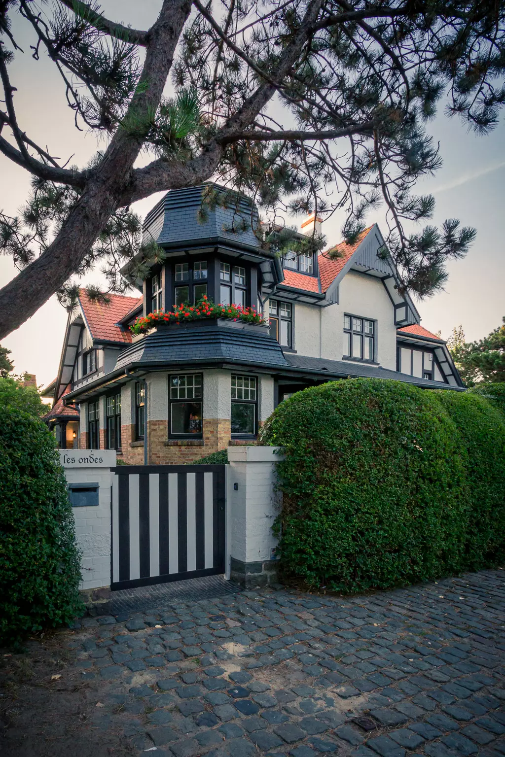 Ein elegantes, mehrstöckiges Haus mit einem markanten Turm, umgeben von grünen Büschen und einem gemusterten Holztor.