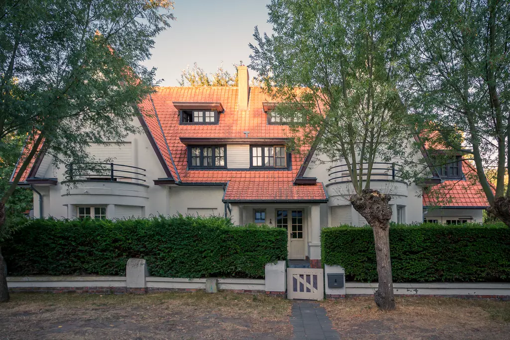 Ein großes, helles Haus mit rotem Satteldach, umgeben von grünen Büschen und Bäumen, vor einem gepflasterten Gehweg.