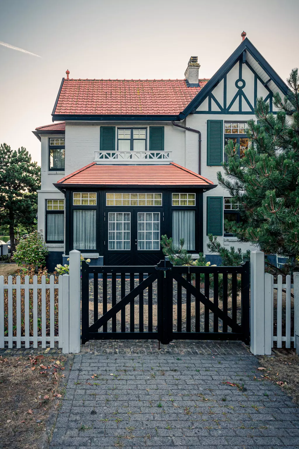 Ein zweistöckiges, klassisches Haus mit rotem Satteldach, weißen Wänden, grünen Fensterläden und einem schwarzen Holzgitterzaun.