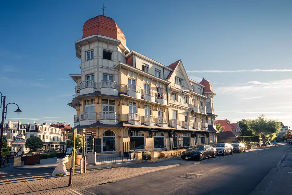 Ein elegantes Gebäude mit einem roten Dach, Balkonen und einer geschwungenen Fassade, umgeben von Bäumen und geparkten Autos.