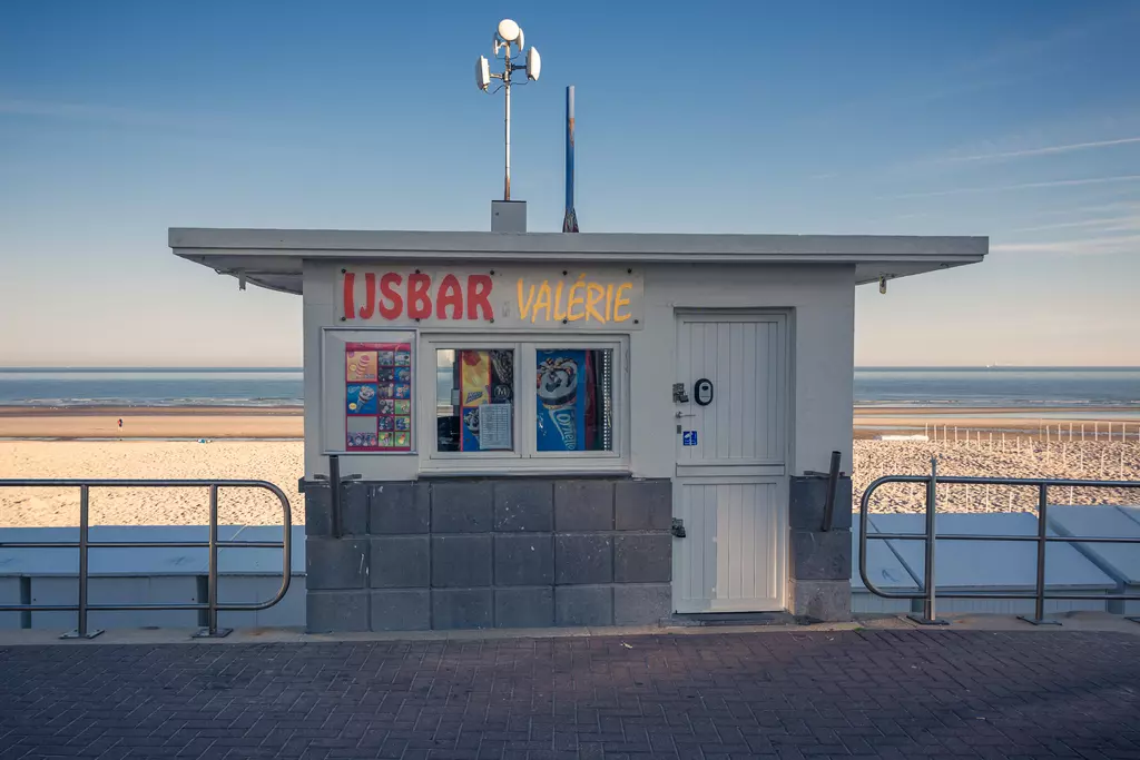 Eine Eisdiele mit einem hellen, einladenden Kiosk, der am Strand steht, umgeben von sandigem Bereich und einem klaren Himmel.