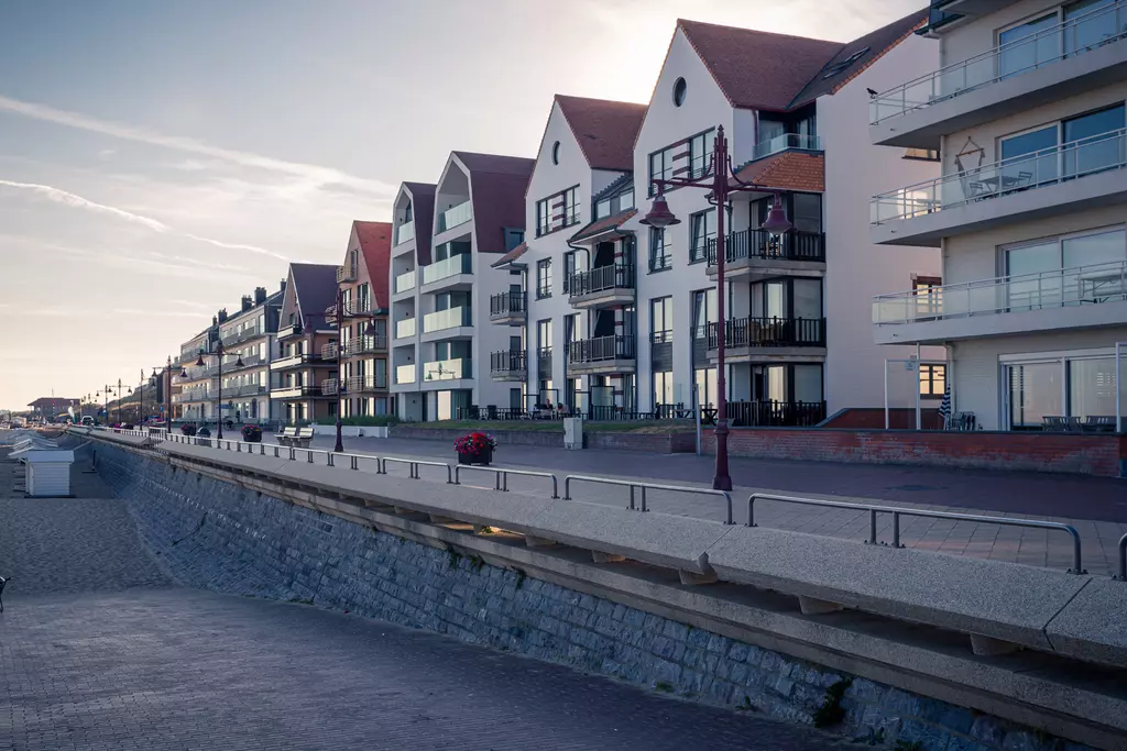 Mehrstöckige Gebäude in hellen Farben, die entlang der Küstenpromenade stehen, mit Bäumen, Laternen und einem Kiesweg im Vordergrund.