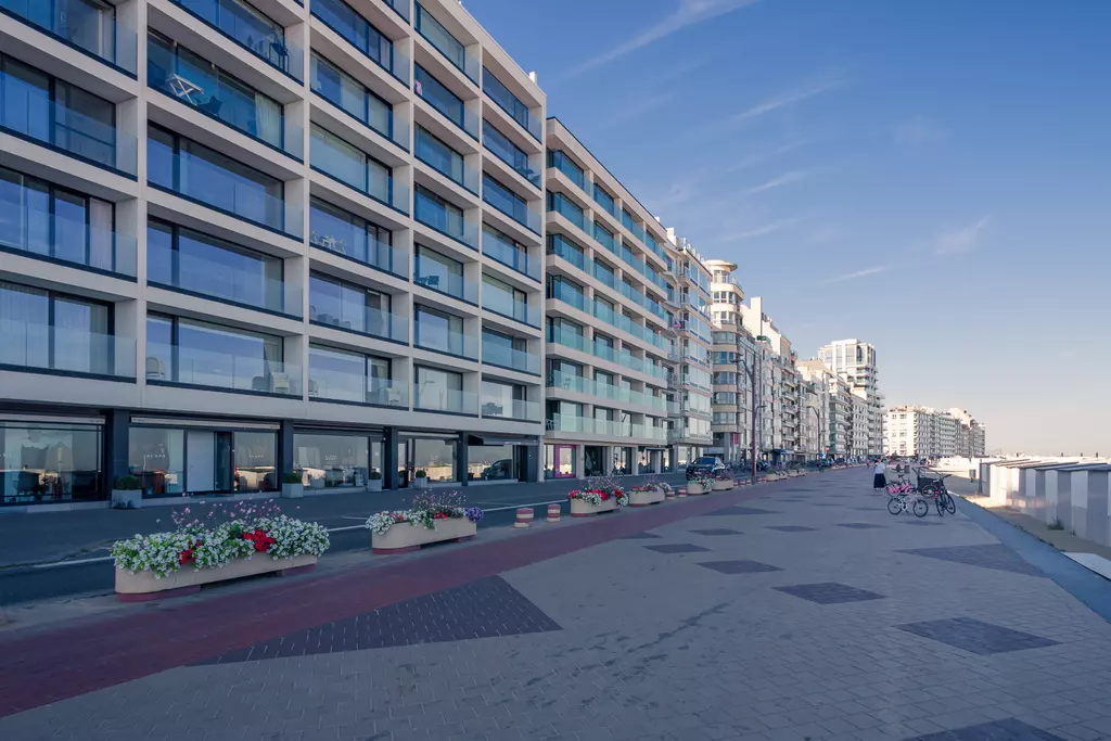 View of modern apartment buildings lining a broad promenade with flower beds and cyclists on the path.