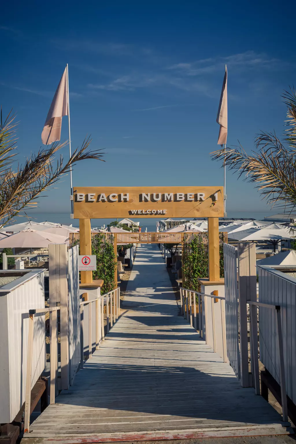 A wooden walkway leads to a sign reading Beach Number 1 and Welcome, flanked by sun umbrellas and palm trees.