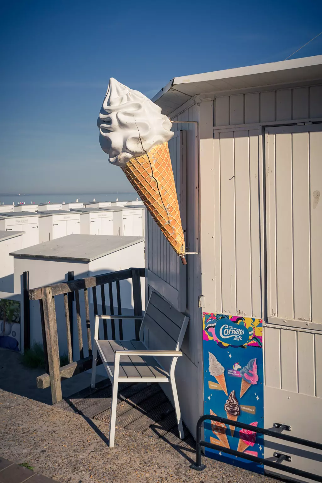 A large, decorative waffle cone hangs from a beach hut. An empty chair is next to it, with an ice cream sign on the wall.