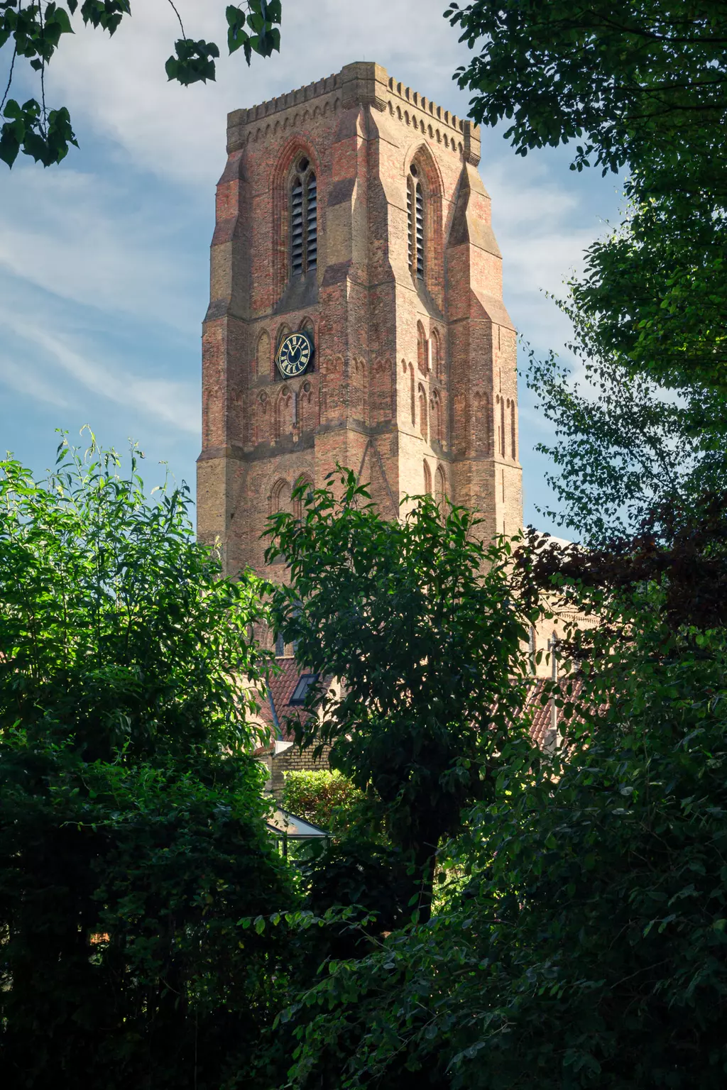 Ein hoher Backsteinturm mit Uhr, umgeben von grünen Bäumen und Sträuchern, unter einem blauen Himmel mit leichten Wolken.