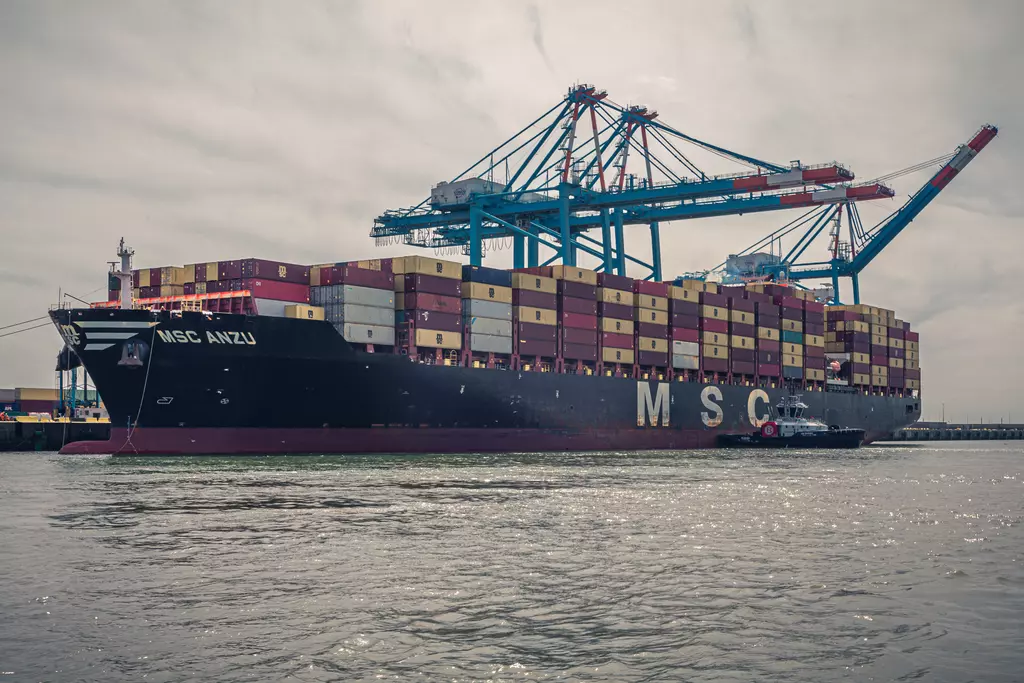 Großes Containerschiff mit verschiedenen Containerfarben und Hafenkränen im Hintergrund, reflektiert im Wasser.