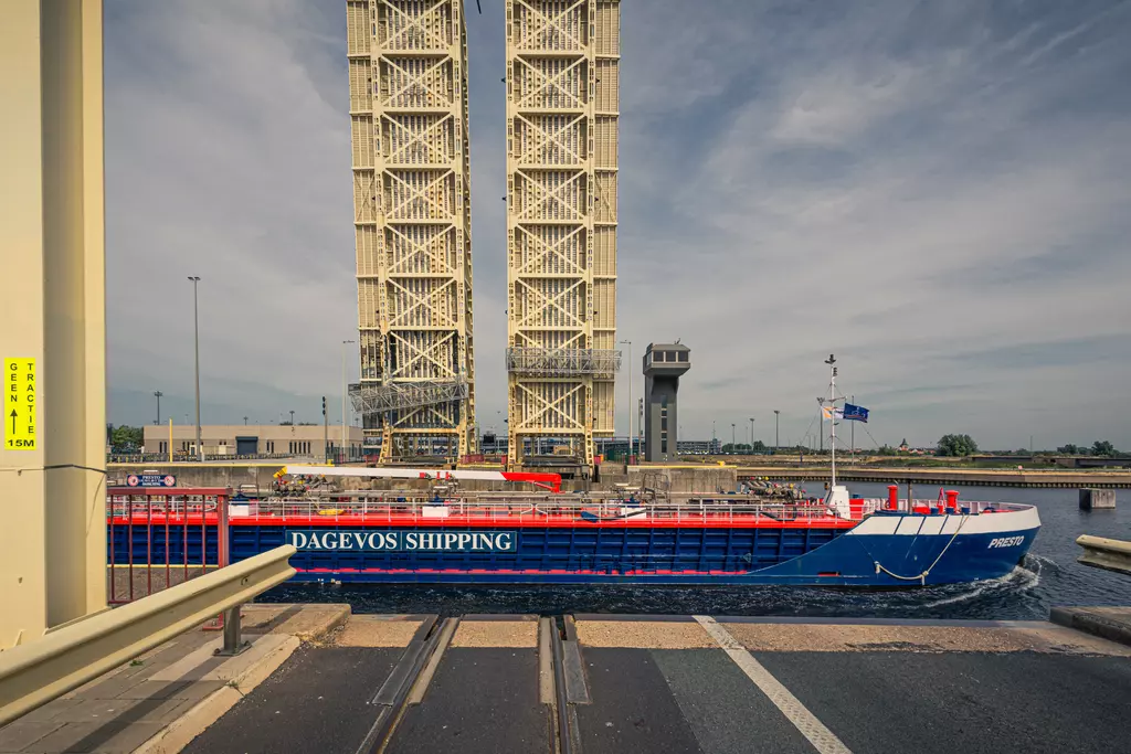 Ein Frachtschiff fährt unter einer geöffneten Klappbrücke hindurch, im Hintergrund sind Hafenanlagen und ein Kontrollturm sichtbar.