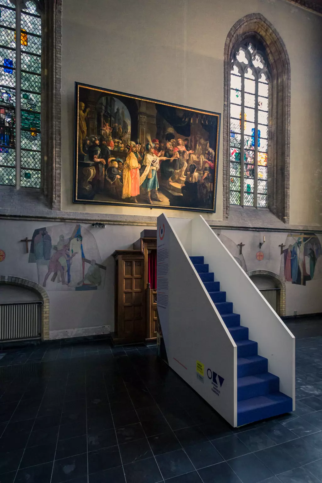 Inside a space featuring a large wall painting and colorful windows, a white staircase with blue steps is visible.