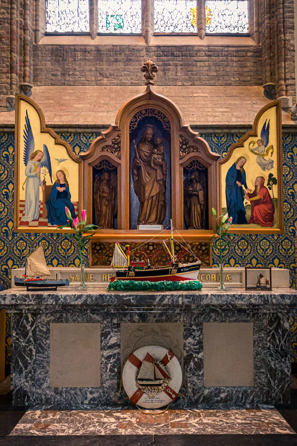 A wooden altar featuring a statue and images of religious figures, flanked by a model ship and a stone plaque.