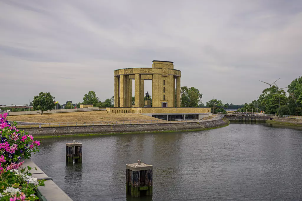 A monumental structure with columns stands by a calm riverside, surrounded by trees and colorful flowers.