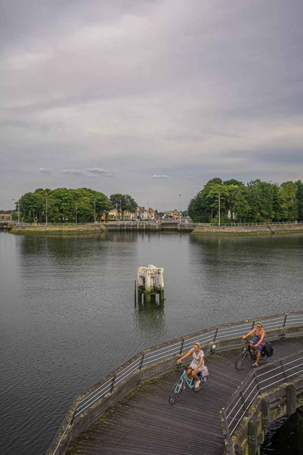 Two cyclists on a wooden promenade by a calm body of water, surrounded by trees and a small town in the background.