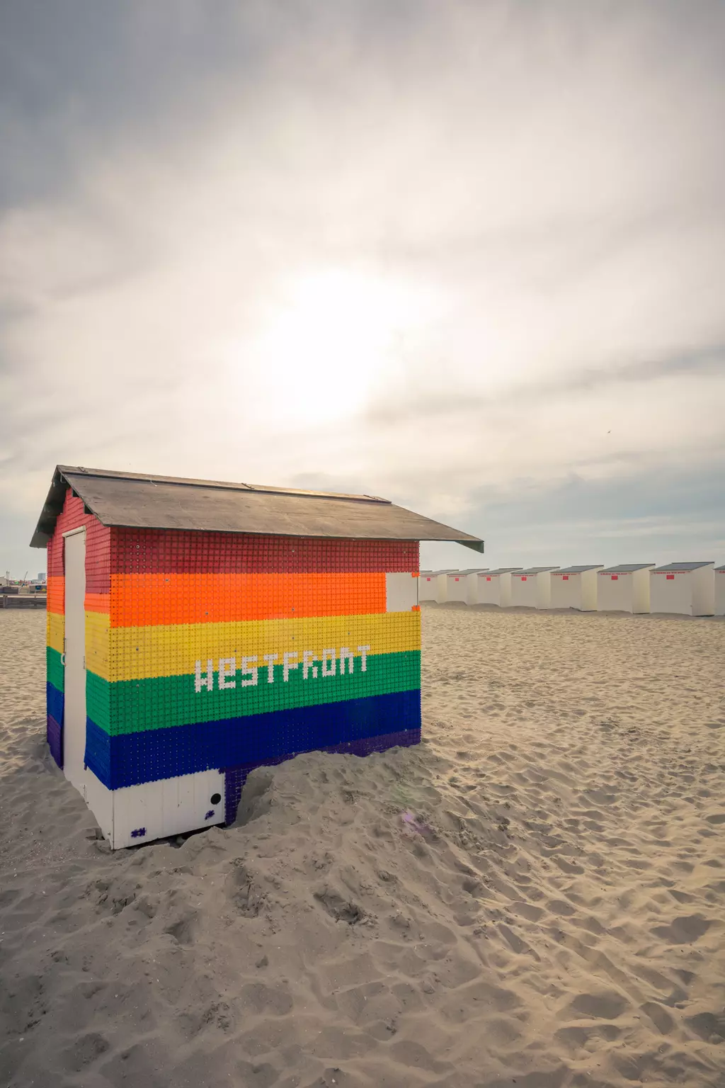 A colorful beach hut in rainbow colors stands on the sand, with additional huts visible in the background.