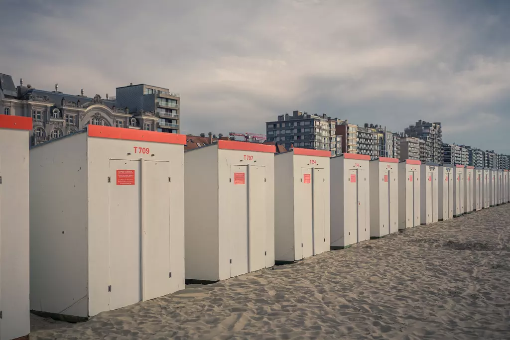 A row of white beach cabins with red roofs sits on sandy ground, with apartment buildings and a cloudy sky in the background.