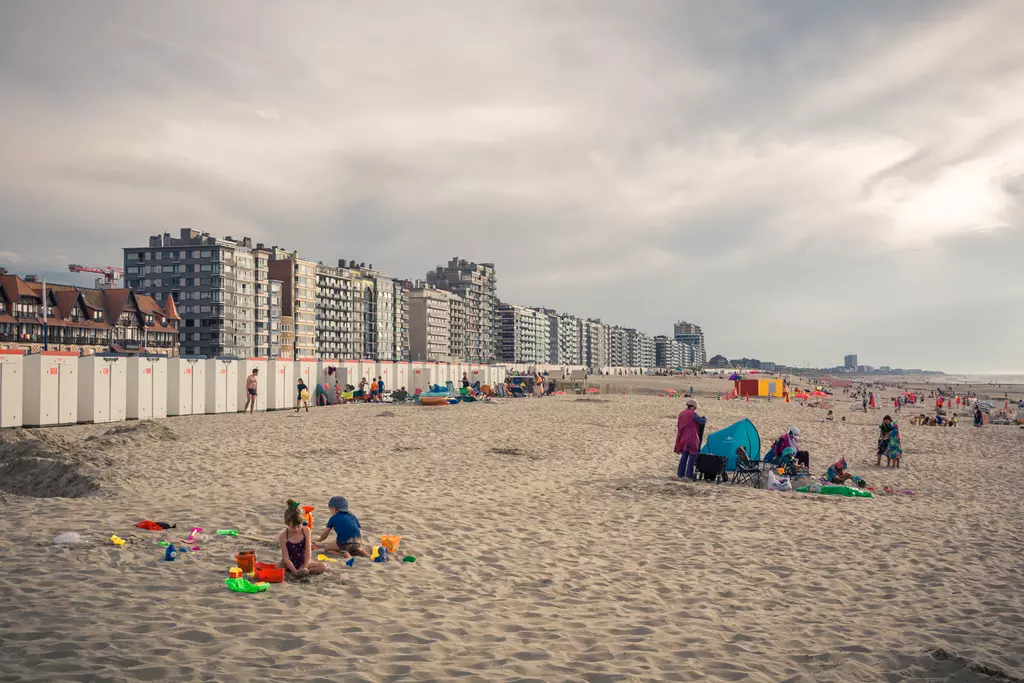 People play in the sand at the beach, surrounded by buildings and beach huts, with a cloudy sky overhead.