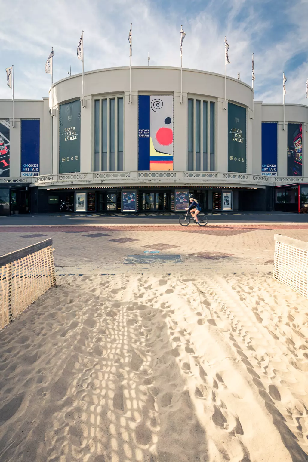 A view of a modern building with flags, colorful posters, and a sandy area in the foreground. A cyclist is passing by.