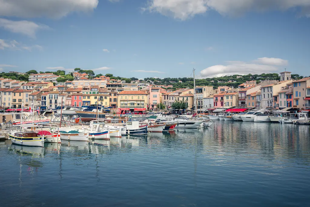 Hafen von Cassis mit mehreren Booten im Wasser und bunten Gebäuden am Ufer. Im Hintergrund ist eine grüne Hügellandschaft zu sehen.