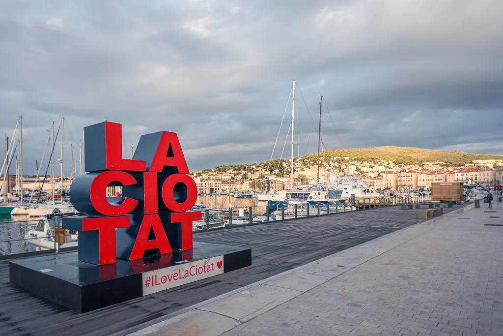 Colorful "LA CIOTAT" sign at the marina in La Ciotat, with boats and hills in the background against a cloudy sky.