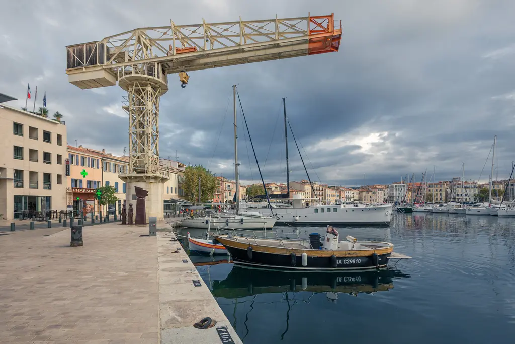 A scenic view of La Ciotat harbor featuring a crane, moored boats, and charming waterfront buildings under a cloudy sky.