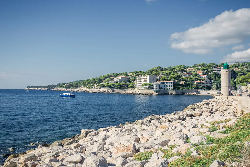 Küstenansicht von Cassis mit felsiger Uferlinie, einem Boot im Wasser und grünen Hügeln sowie Gebäuden im Hintergrund.