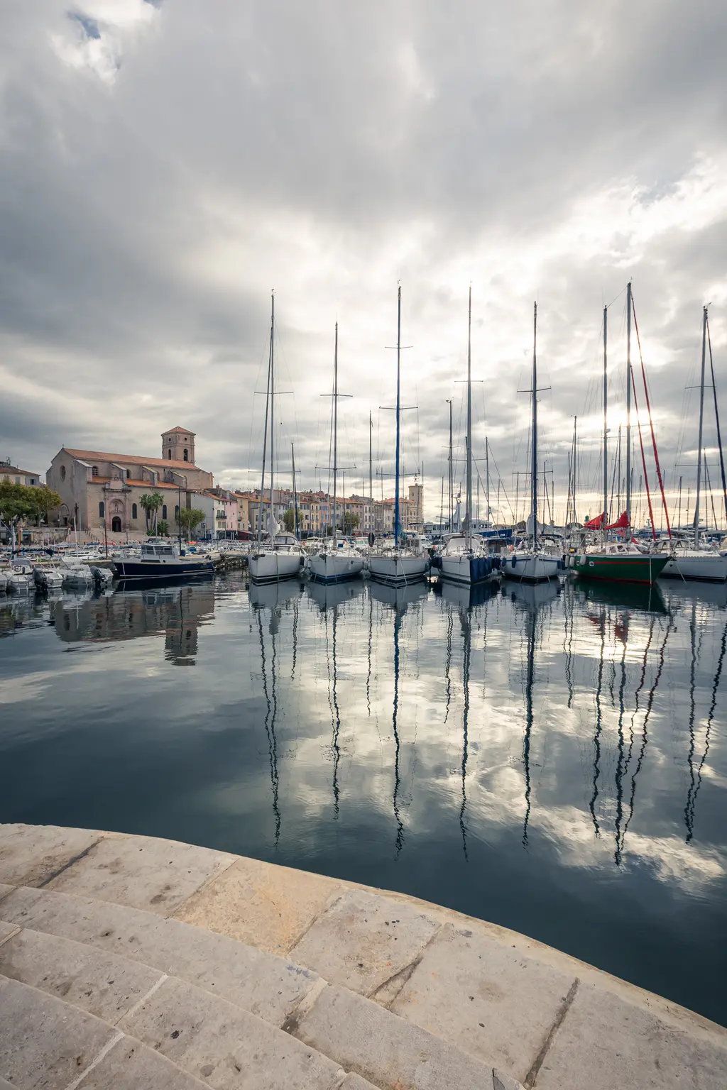 A marina in La Ciotat with sailboats reflecting in calm water under a cloudy sky, featuring a historic building in the background.