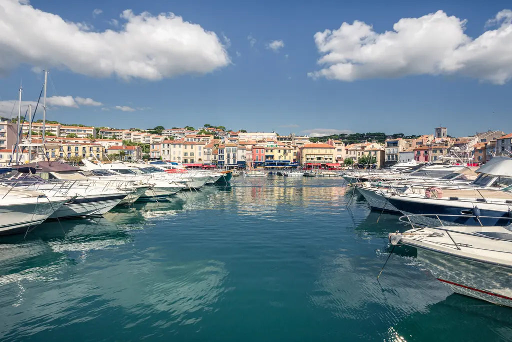 Hafen von Cassis mit zahlreichen Booten im klaren Wasser. Im Hintergrund stehen bunte Häuser und ein blauer Himmel mit einigen Wolken.