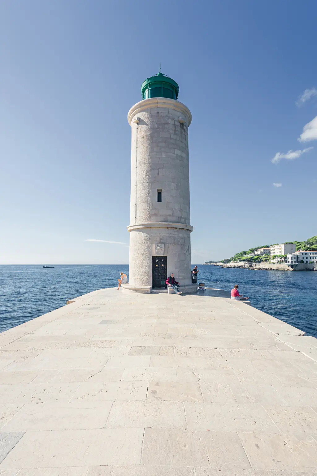 Leuchtturm in Cassis auf einem Steg, umgeben von Wasser. Im Hintergrund sind Bauten an der Küste und weitere Personen zu sehen.