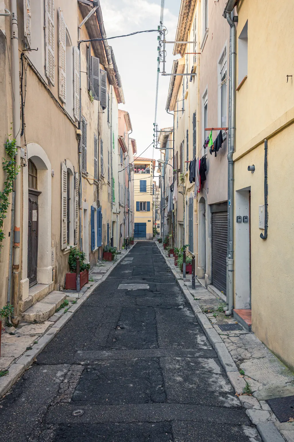 A narrow street in La Ciotat lined with rustic buildings, blue window shutters, and potted plants on either side.