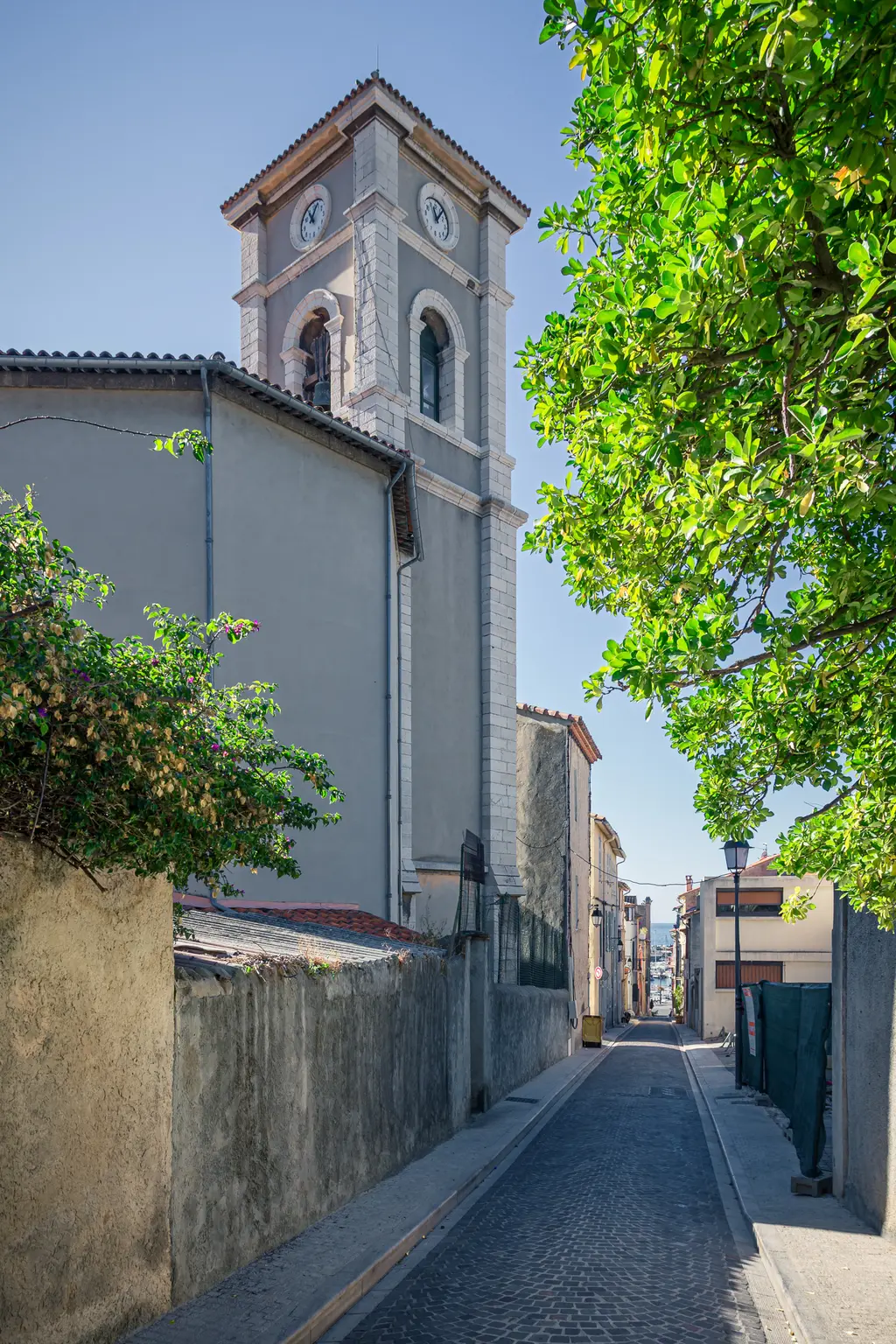 Straße in Cassis mit der Église Saint-Michel im Hintergrund. Die schmalen Häuser flankieren die gepflasterte Straße, umgeben von Bäumen.