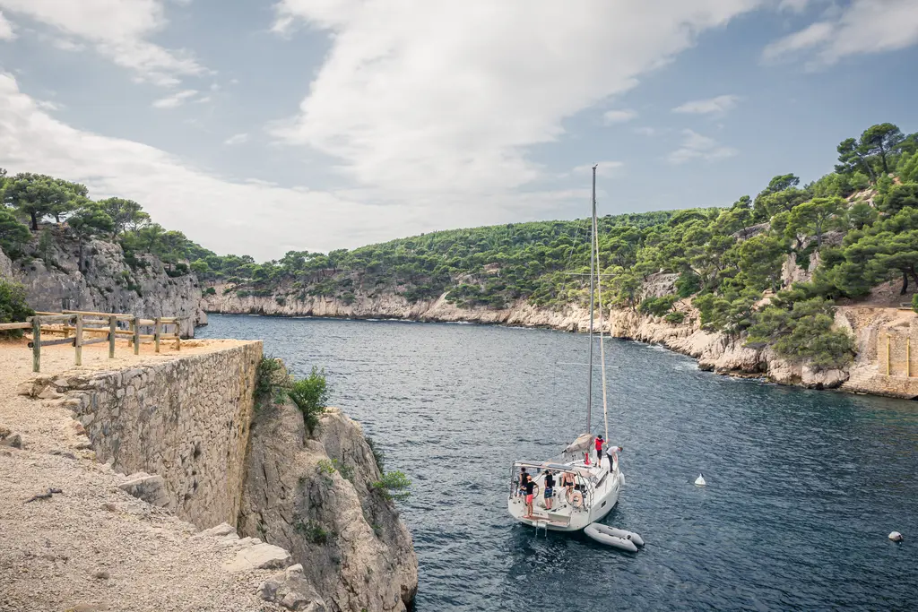 Bucht in Cassis mit einem Segelboot im Wasser, umgeben von steilen Felsen und grünen Bäumen. Der Himmel ist bewölkt.