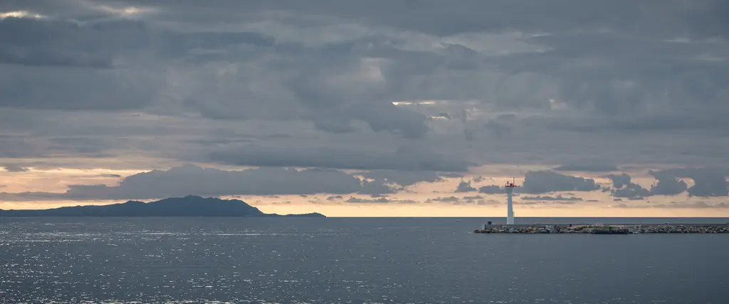 A lighthouse in La Ciotat stands on a rocky pier, surrounded by calm waters and dramatic clouds in the sky.