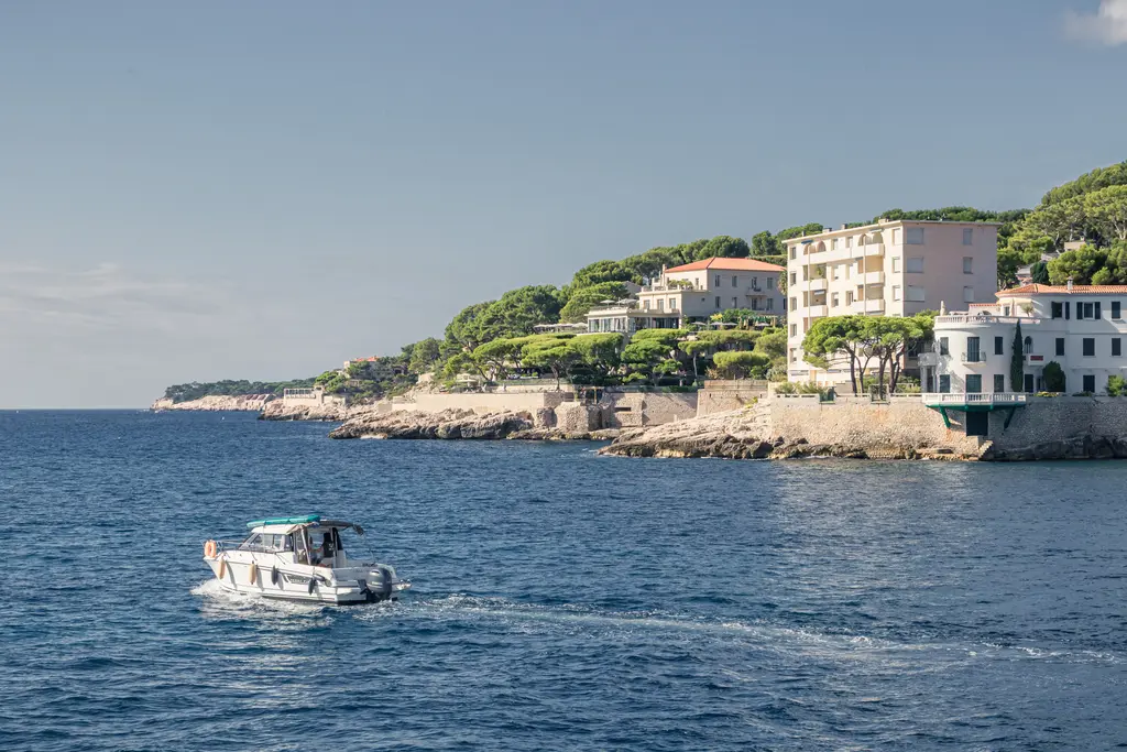 Küstenansicht von Cassis mit einem Boot, das auf dem blauen Wasser fährt, umgeben von eleganten Gebäuden und grünen Bäumen.