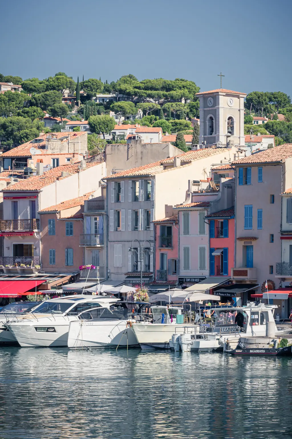 Bunte Häuser im Hafen von Cassis mit einem Kirchturm im Hintergrund. Mehrere Boote liegen am Ufer, das Wasser spiegelt die Gebäude wider.