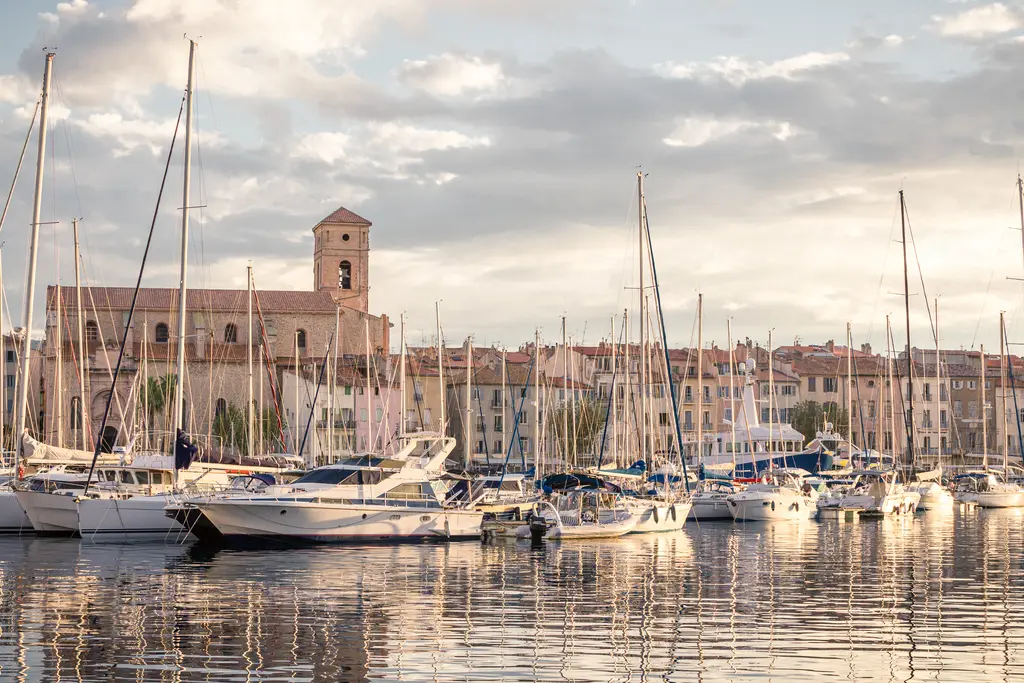 A marina in La Ciotat with sailboats reflected in calm waters and historic buildings in the background. Soft evening light enhances the scene.