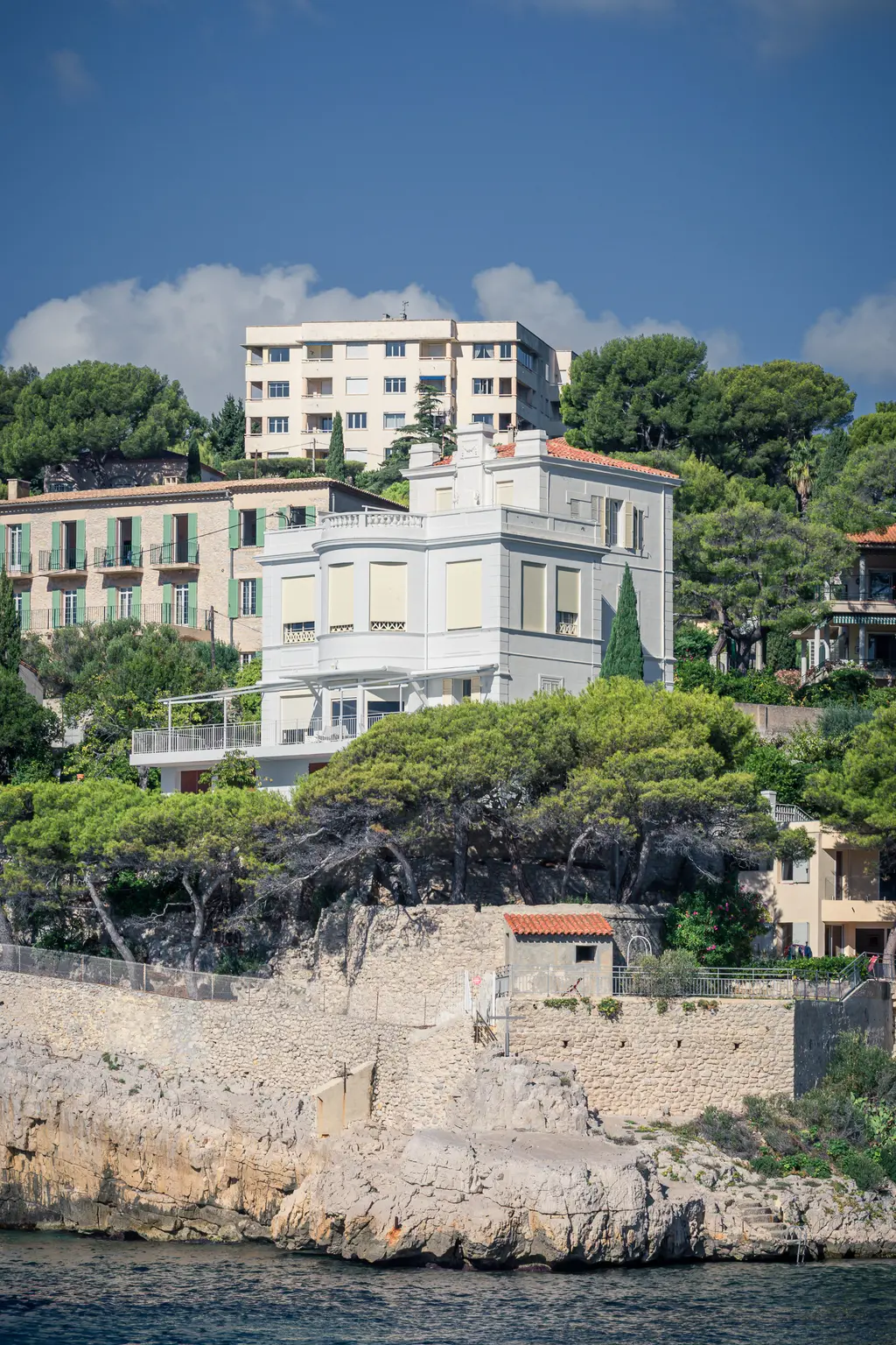 Hübsches Wohnhaus in Cassis, umgeben von Bäumen, auf einer Klippe über dem Wasser. Im Hintergrund mehrere Gebäude und ein blauer Himmel.