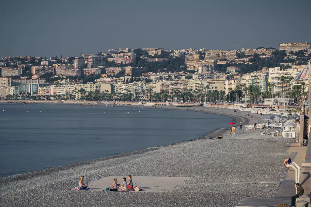 Gray pebble beaches with a few people relaxing, while city architecture and palm trees are visible in the background.