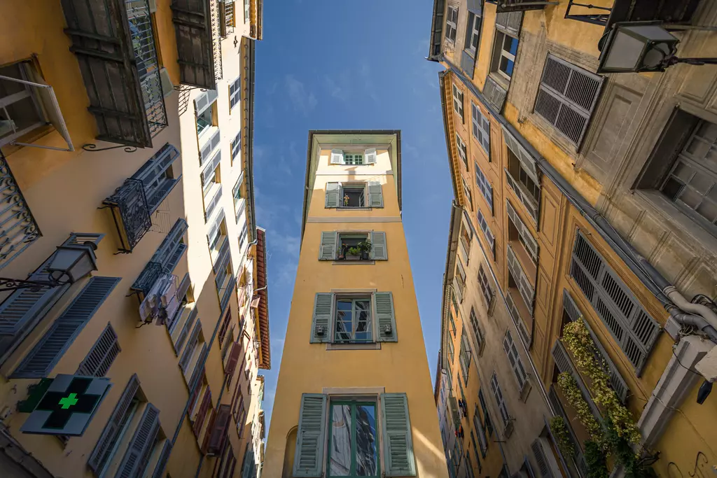 A narrow yellow tower stands between colorful, multi-story buildings. The sky is clear and blue, with windows visible.