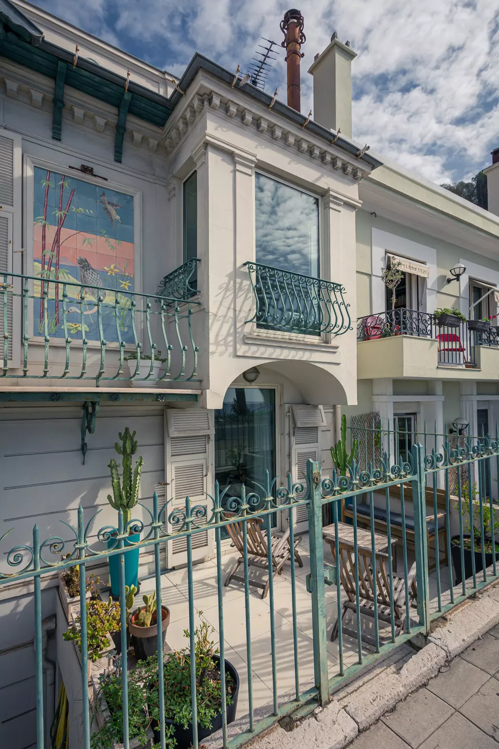 A bright building facade with green balcony railings, plants, and a table on the terrace under a cloudy sky.