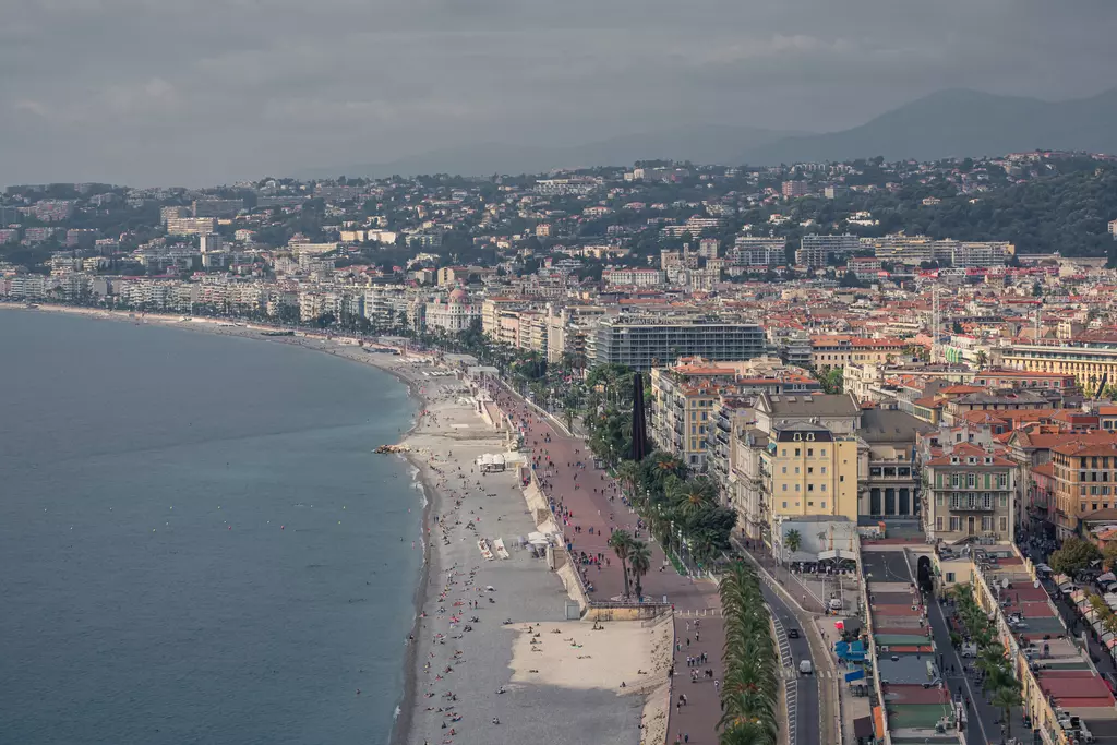 A coastal view featuring a long promenade, sandy beach, and buildings. The sea glistens in various shades of blue under a cloudy sky.