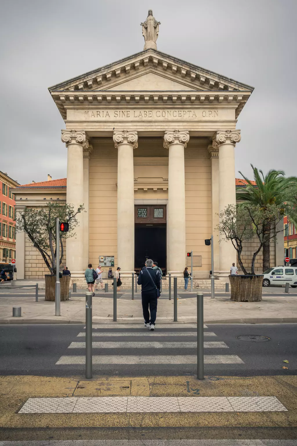 A neoclassical building with columns and a statue on the roof, surrounded by pedestrians and palm trees.