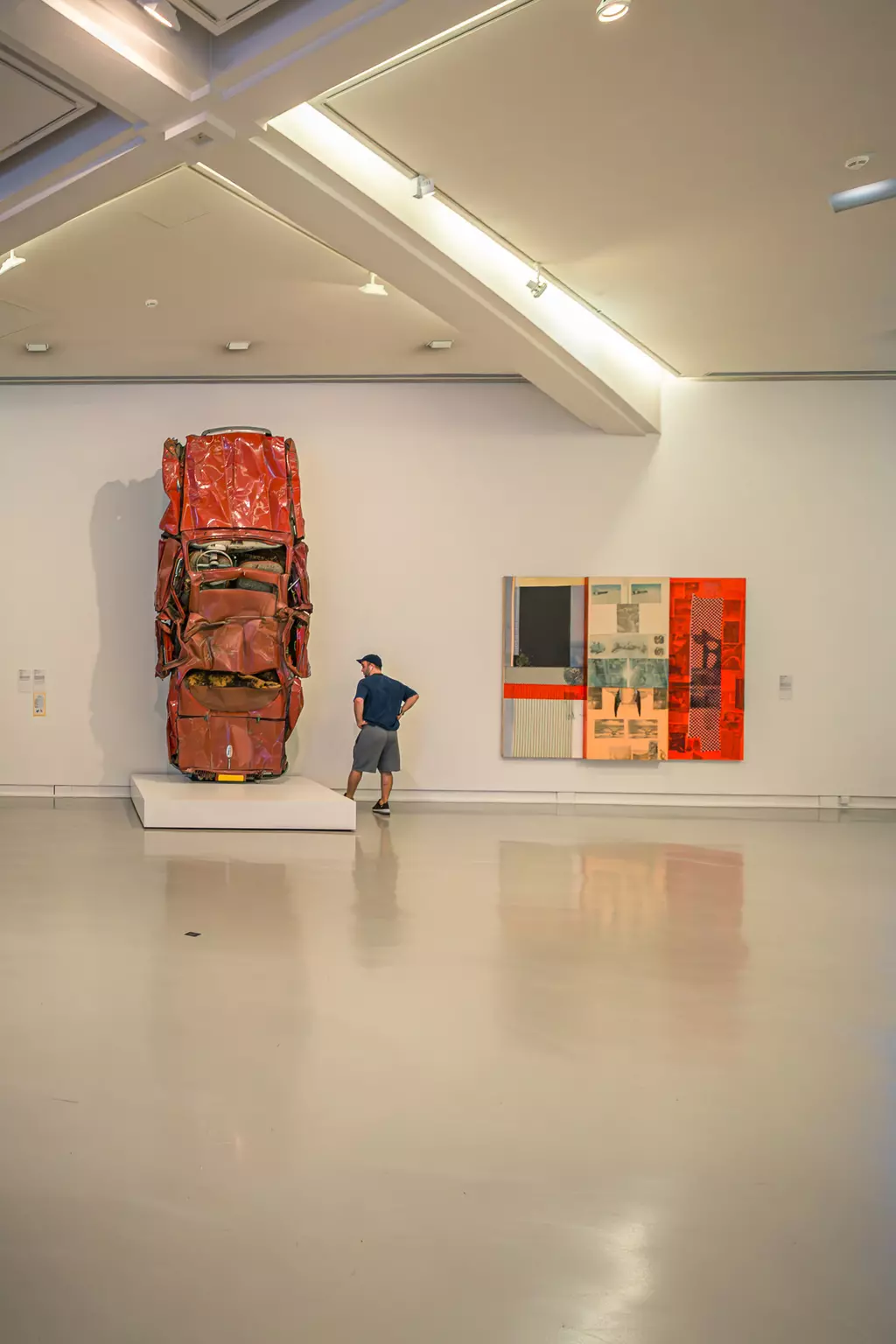 A man stands in a modern gallery in front of a large red sculpture on a pedestal, with colorful artworks on the right.