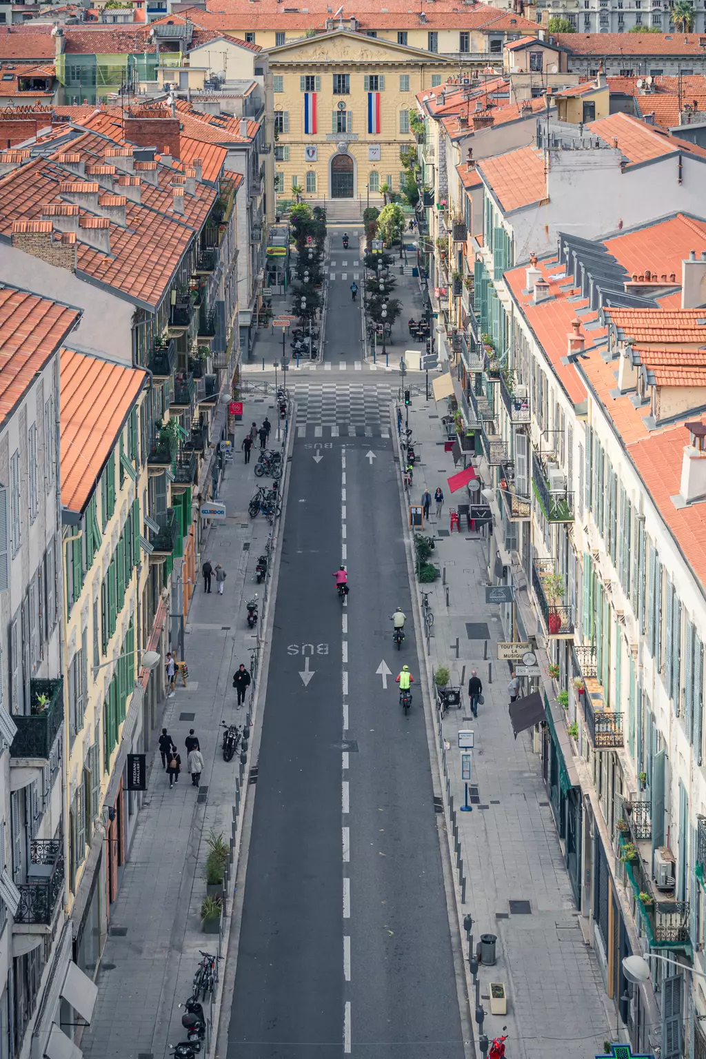 A narrow street with cyclists and pedestrians, lined with buildings with red roofs, leads to a yellow historic building.