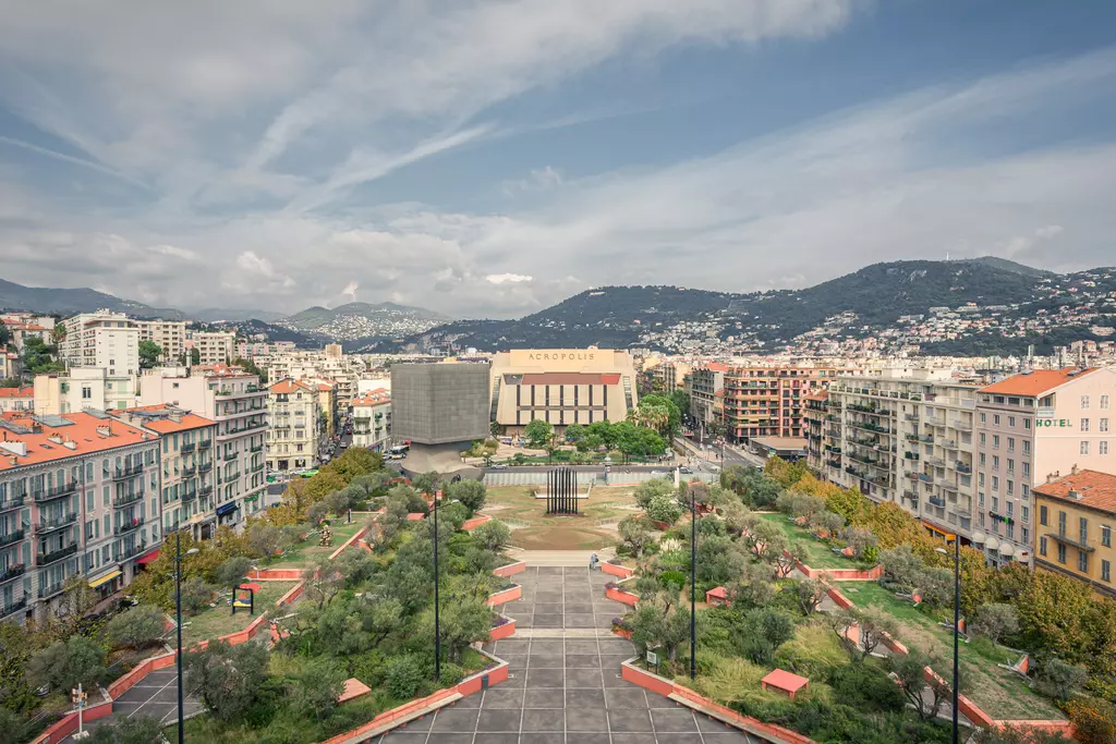 A view of an urban landscape with modern buildings, a park with trees, and a hill in the background under a cloudy sky.