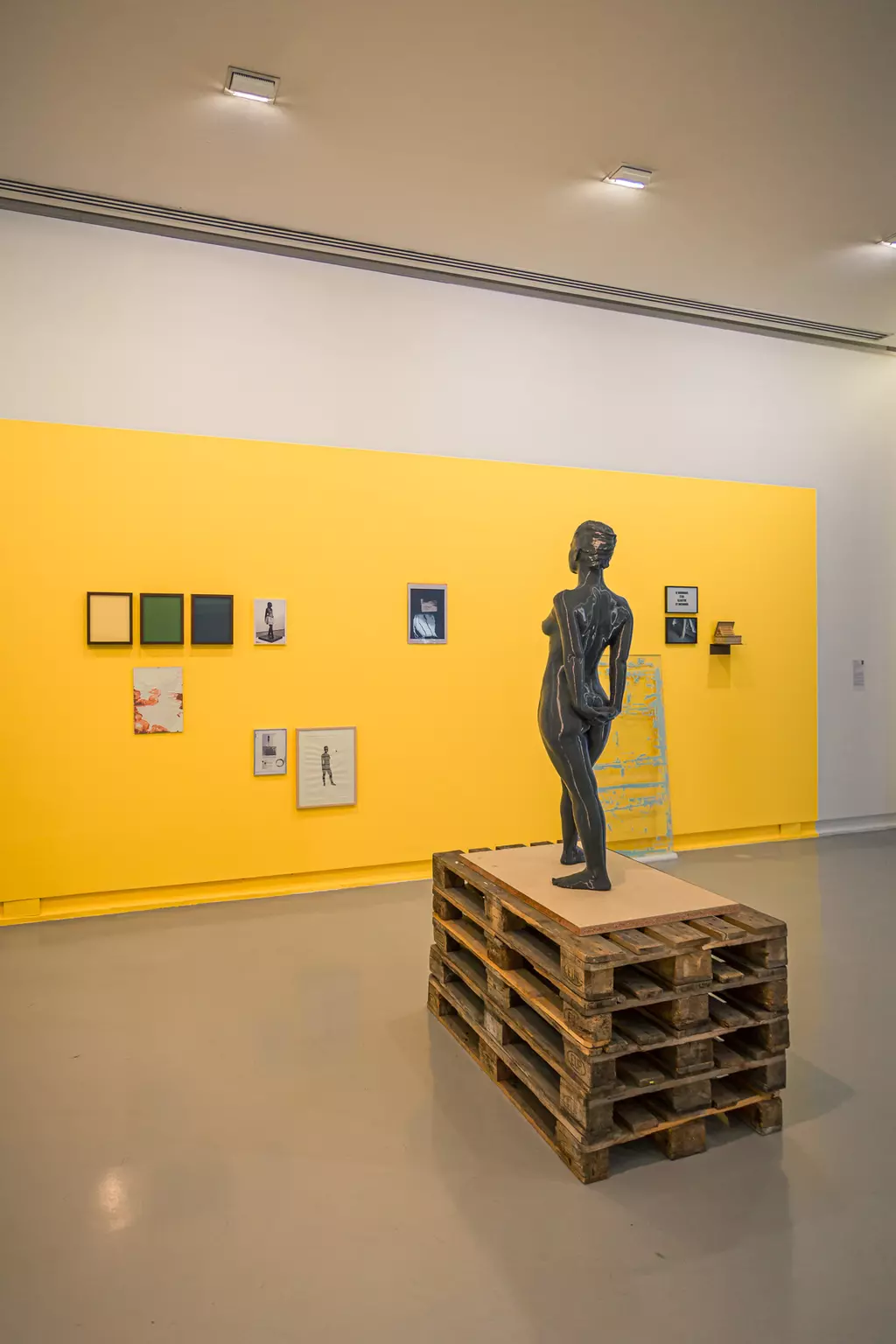 A black statue stands on a wooden palette against a yellow wall, surrounded by various framed artworks.