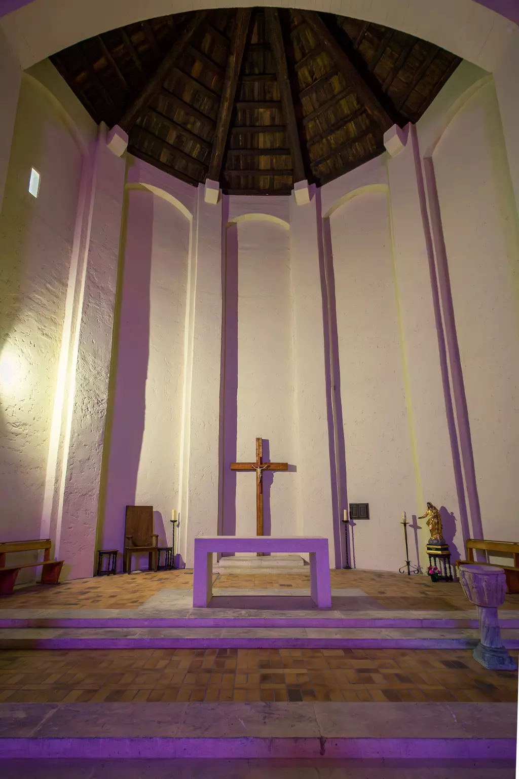 Ein rechteckiger Raum mit einer Holzdecke, einem altären Tisch, einem Kreuz an der Wand und bunter Beleuchtung.